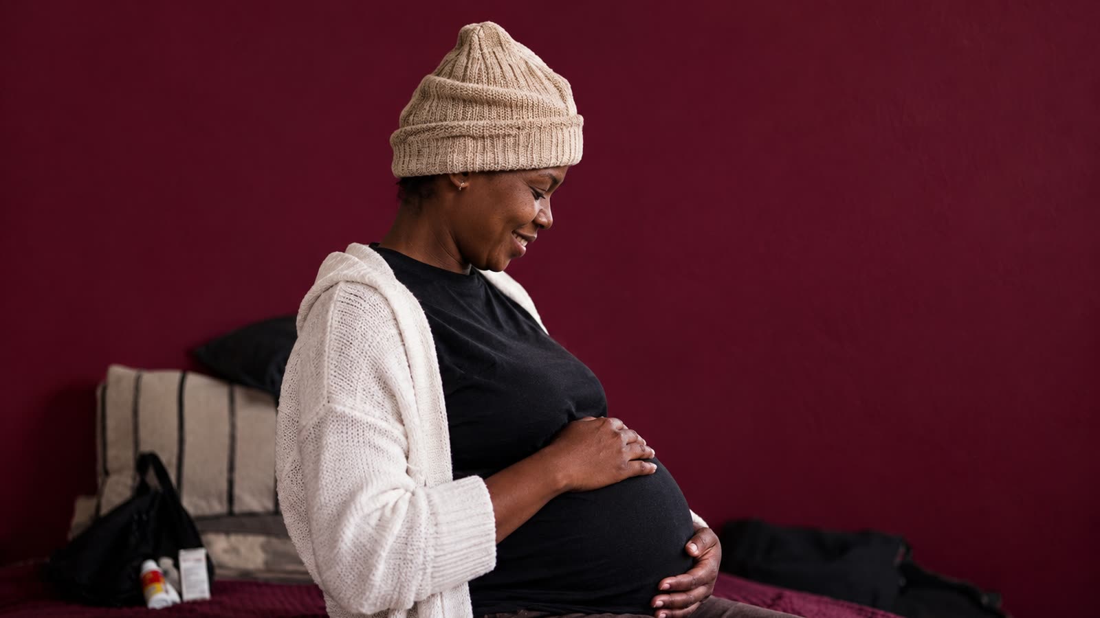 A smiling pregnant woman in a knitted beanie sitting on a bed, holding her bump